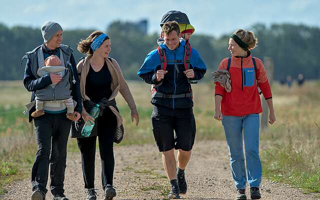 <p>Zwei Familien mit Kleinkindern in Tragerucksäcken wandern über ein Feld.</p>